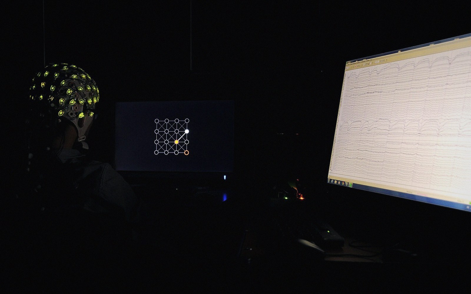 A person wearing an EEG electrode cap faces a screen showing a node diagram beside a brainwave readout monitor