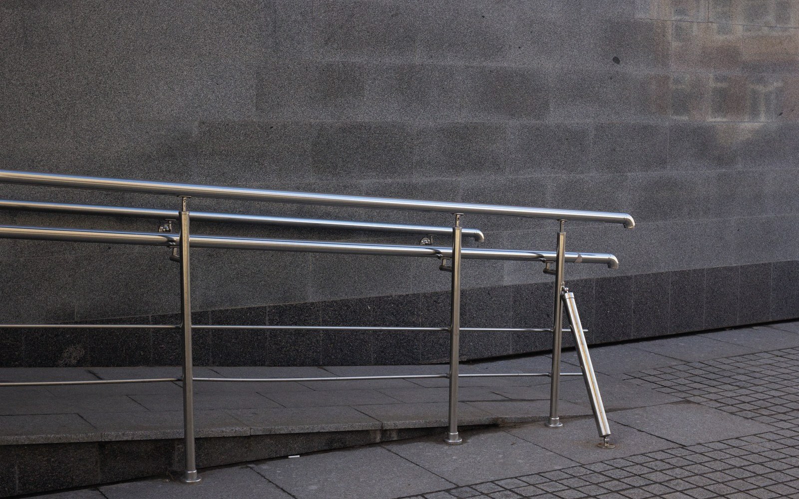 Stainless steel handrails line a stone ramp beside a granite wall